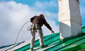 Pracownik na dachu, zabezpieczony linami, wykonuje malowanie chlorokauczukiem zielonego dachu i komina.