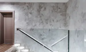 Nowoczesny przedpokój ze schodami i szklaną balustradą. Ściany pokryte szarym tynkiem strukturalnym, który świetnie sprawdzi się w tym miejscu.