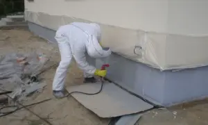 Pracownik w kombinezonie ochronnym wykonuje malowanie elewacji metodą natryskową, zabezpieczając teren folią.