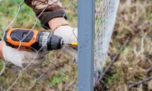 Ręce w rękawiczkach przykręcają siatkę do słupka. Widać wiertarkę i elementy ogrodzenia.