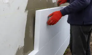 Pracownik w czerwonych rękawicach montuje płyty styropianowe na ścianie. To etap prac związanych z tym, ile kosztuje ocieplenie domu.
