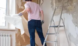 Mężczyzna w rękawiczkach zrywa starą tapetę ze ściany, przygotowując pokój do remontu. Obok stoi drabina.