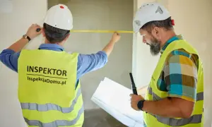 Dwóch inspektorów w kaskach i kamizelkach mierzy pomieszczenie taśmą, jeden trzyma plany.