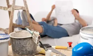 Przerwa w remoncie. Na pierwszym planie wiadro z farbą emulsyjną o spoiwie organicznym, pędzle, młotek i niebieski kask. W tle mężczyzna czyta plany.