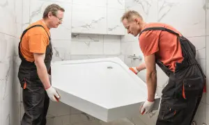 Dwóch fachowców montuje brodzik akrylowy bez nóżek. Prace w łazience z białymi płytkami.