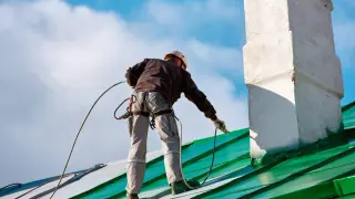 Pracownik na dachu, zabezpieczony linami, wykonuje malowanie chlorokauczukiem zielonego dachu i komina.