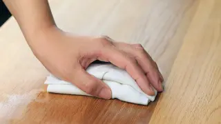 Dłoń z białą szmatką wciera olej w surowe drewno sosnowe, nadając mu piękny, ciepły odcień. To świetny sposób, czym pomalować surowe drewno sosnowe.