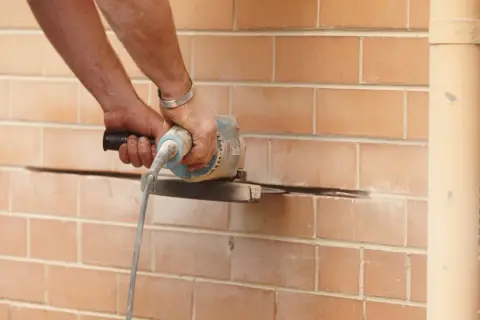 Ręce trzymają szlifierkę kątową, która przecina ceglaną ścianę. Prace remontowe przy nadprożu stalowym.