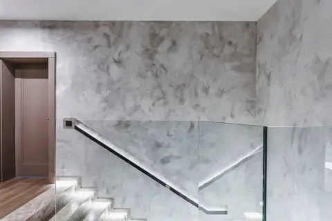 Nowoczesny przedpokój ze schodami i szklaną balustradą. Ściany pokryte szarym tynkiem strukturalnym, który świetnie sprawdzi się w tym miejscu.