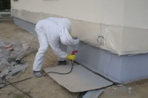 Pracownik w kombinezonie ochronnym wykonuje malowanie elewacji metodą natryskową, zabezpieczając teren folią.