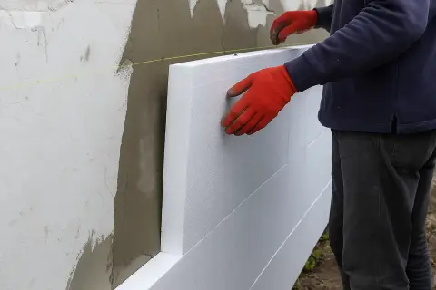 Pracownik w czerwonych rękawicach montuje płyty styropianowe na ścianie. To etap prac związanych z tym, ile kosztuje ocieplenie domu.