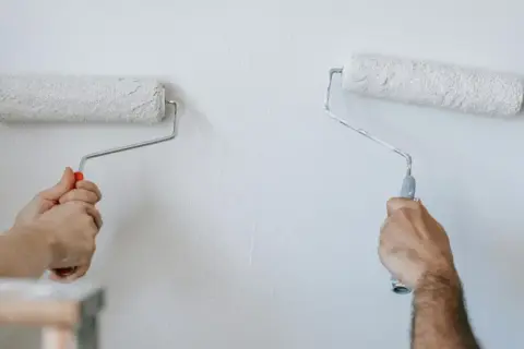 Dwie ręce malują ścianę wałkami. Wybór między farba lateksowa czy akrylowa zależy od efektu.