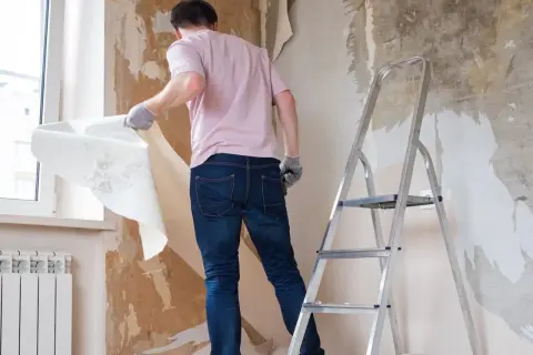 Mężczyzna w rękawiczkach zrywa starą tapetę ze ściany, przygotowując pokój do remontu. Obok stoi drabina.
