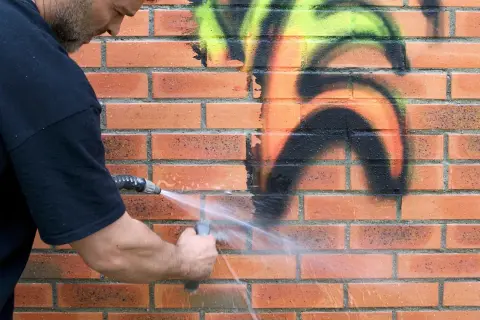 Mężczyzna zmywa graffiti z ceglanej ściany wodą. Dzięki powłoce antygraffiti czyszczenie jest łatwiejsze.