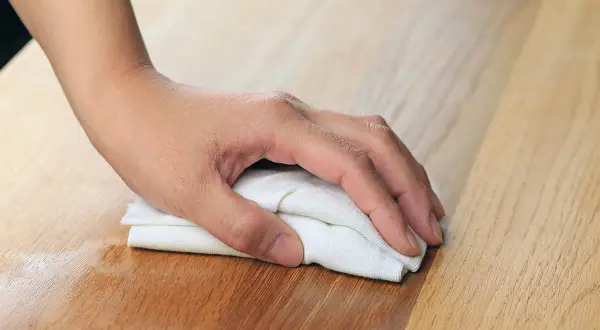 Dłoń z białą szmatką wciera olej w surowe drewno sosnowe, nadając mu piękny, ciepły odcień. To świetny sposób, czym pomalować surowe drewno sosnowe.