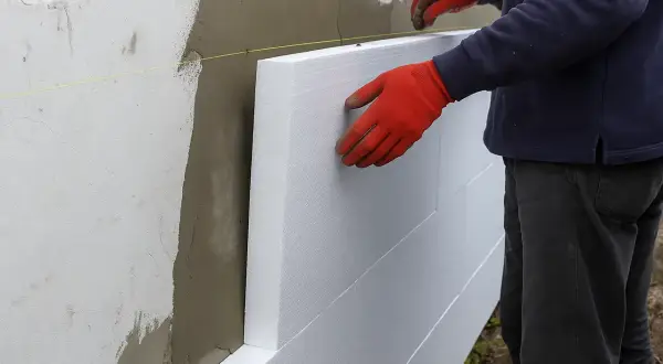 Pracownik w czerwonych rękawicach montuje płyty styropianowe na ścianie. To etap prac związanych z tym, ile kosztuje ocieplenie domu.