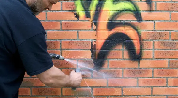 Mężczyzna zmywa graffiti z ceglanej ściany wodą. Dzięki powłoce antygraffiti czyszczenie jest łatwiejsze.