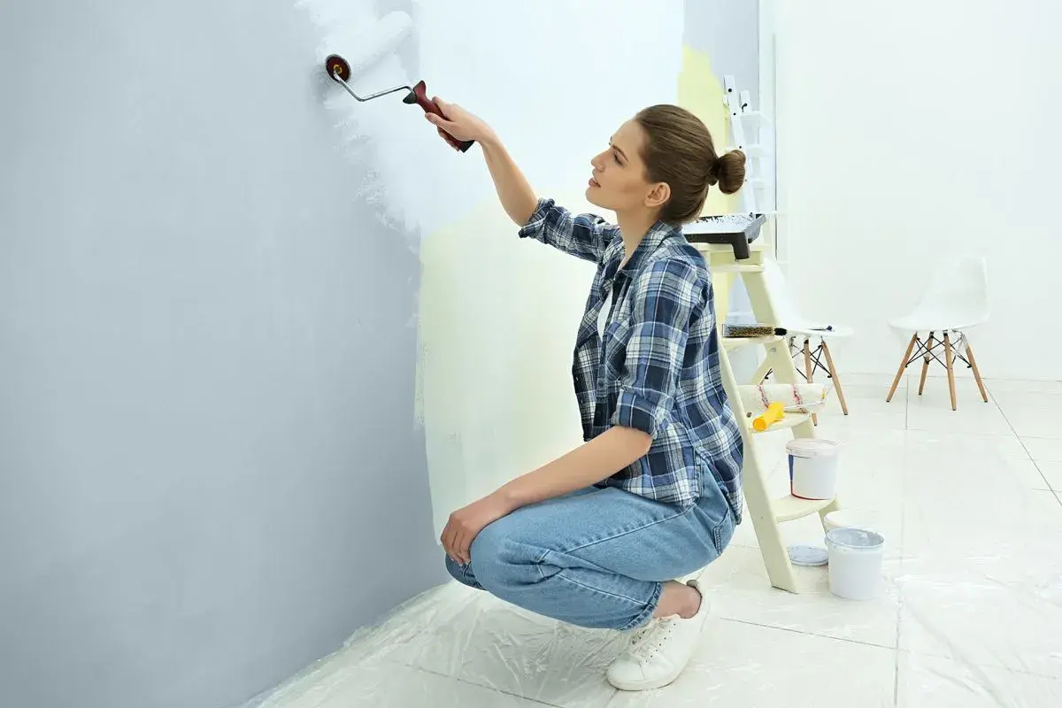Kobieta maluje ścianę wałkiem, dbając o to, jak malować ściany wałkiem bez smug. W tle drabina i wiaderka z farbą.