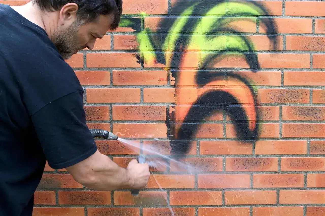 Mężczyzna zmywa graffiti z ceglanej ściany wodą pod ciśnieniem. Dzięki powłoce antygraffiti czyszczenie jest łatwiejsze.