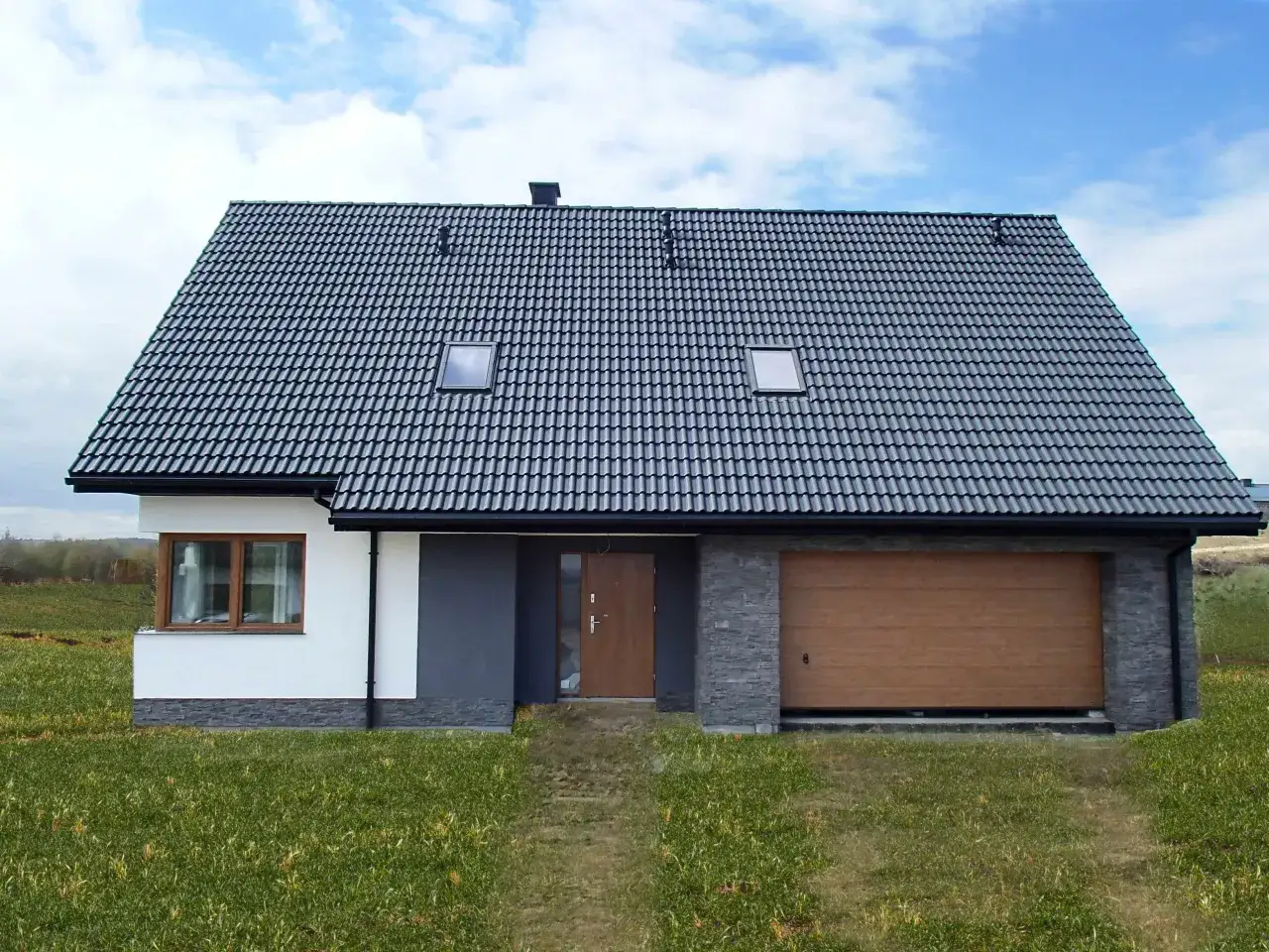 Nowoczesny dom z szarym dachem, brązową bramą garażową i białymi ścianami, na tle błękitnego nieba.