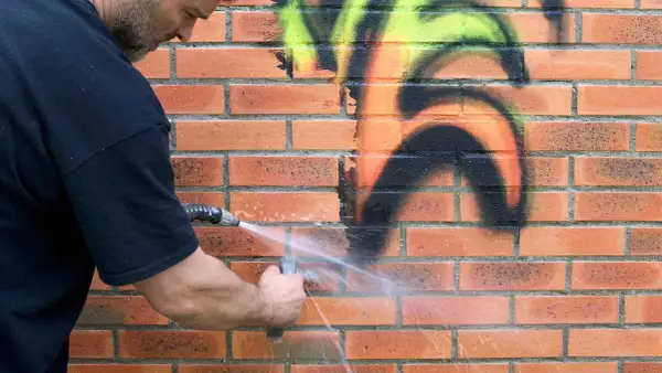 Mężczyzna zmywa graffiti z ceglanej ściany wodą. Dzięki powłoce antygraffiti czyszczenie jest łatwiejsze.